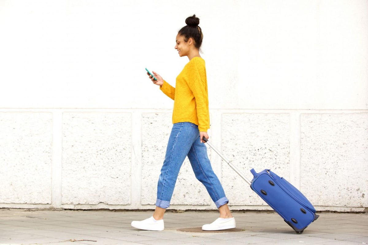 Side view portrait of young woman walking with travel bag and using mobile phone